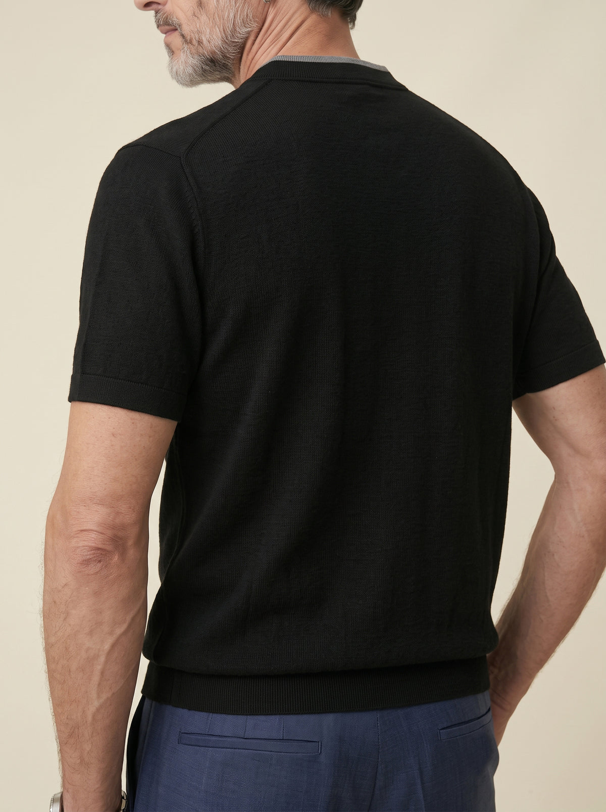 #Jet-Black_group A man wearing a black short-sleeve knit polo shirt, shown from the back against a plain beige background.