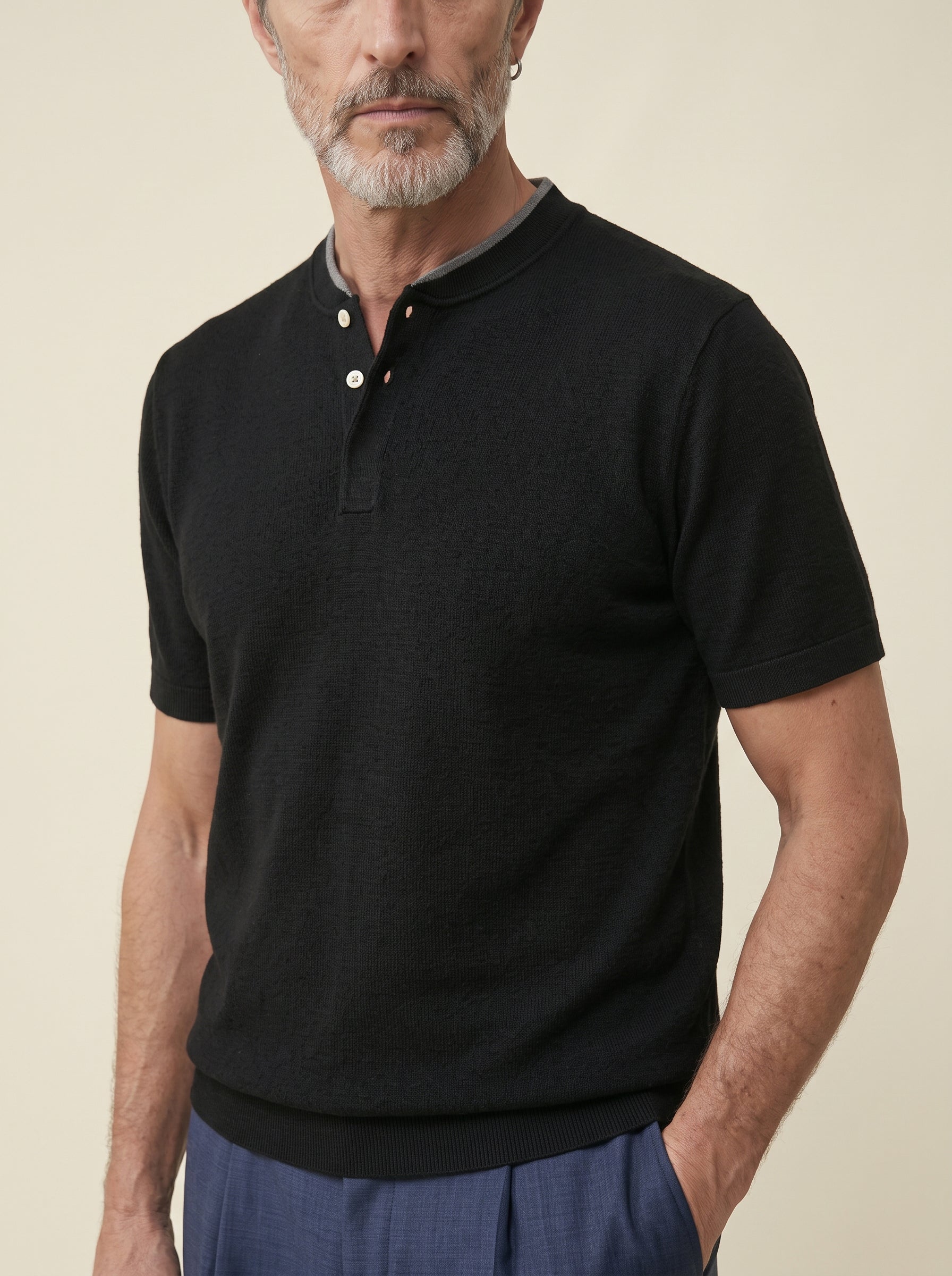#Jet-Black_group A man wearing a black short-sleeve knit polo shirt with a three-button henley collar, paired with blue trousers, against a solid beige background.