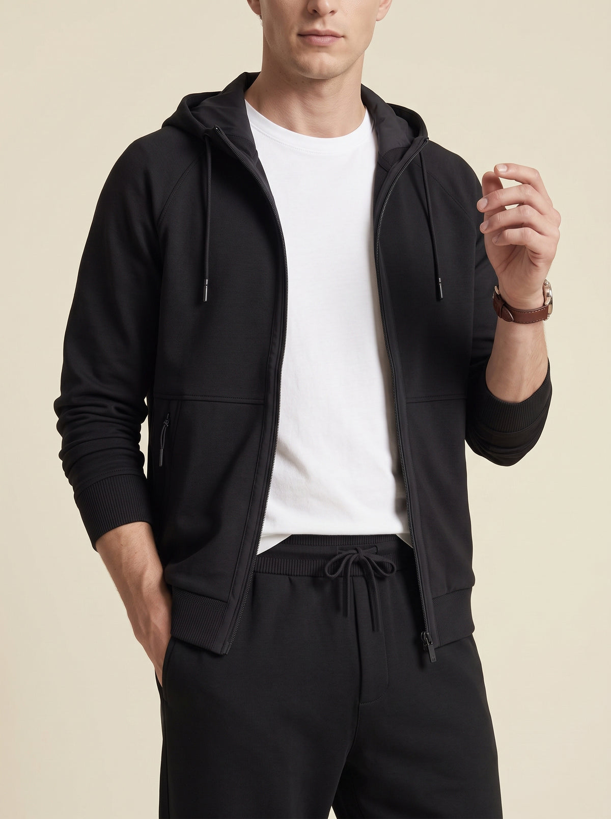 #Jet-Black_group A man wearing a black zip-up hoodie over a white t-shirt and black sweatpants, photographed against a solid light beige background.