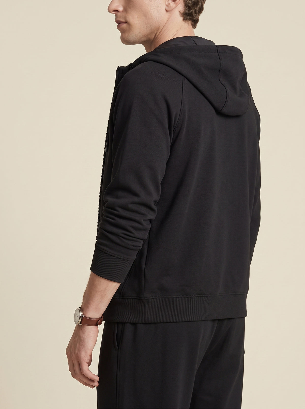 #Jet-Black_group A man wearing a black pullover hoodie and matching black pants, shown from a side-back angle against a plain beige studio background.