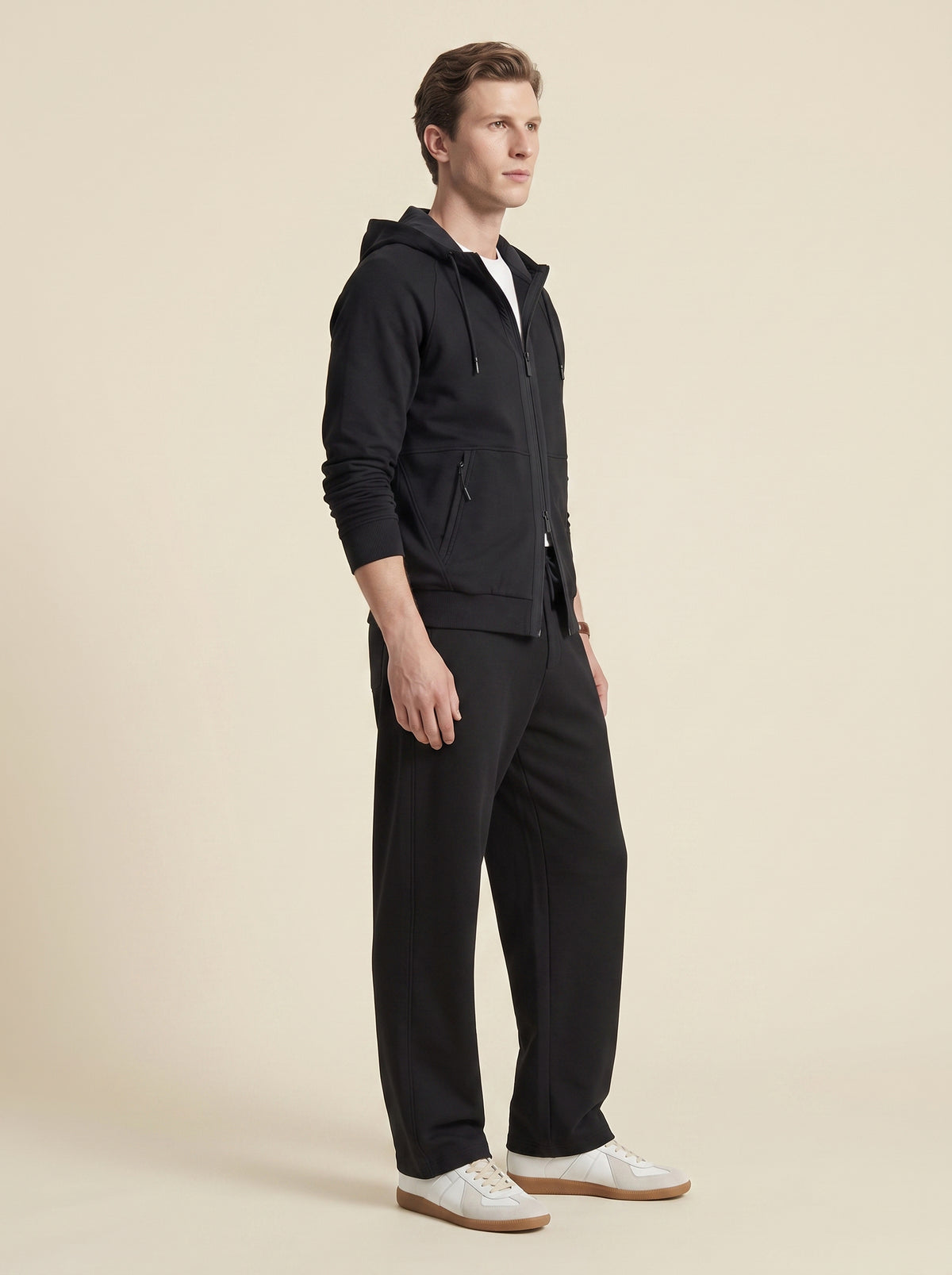 #Jet-Black_group A man wearing a black zip-up hoodie and matching black trousers, paired with white sneakers, standing in profile against a solid beige studio background.