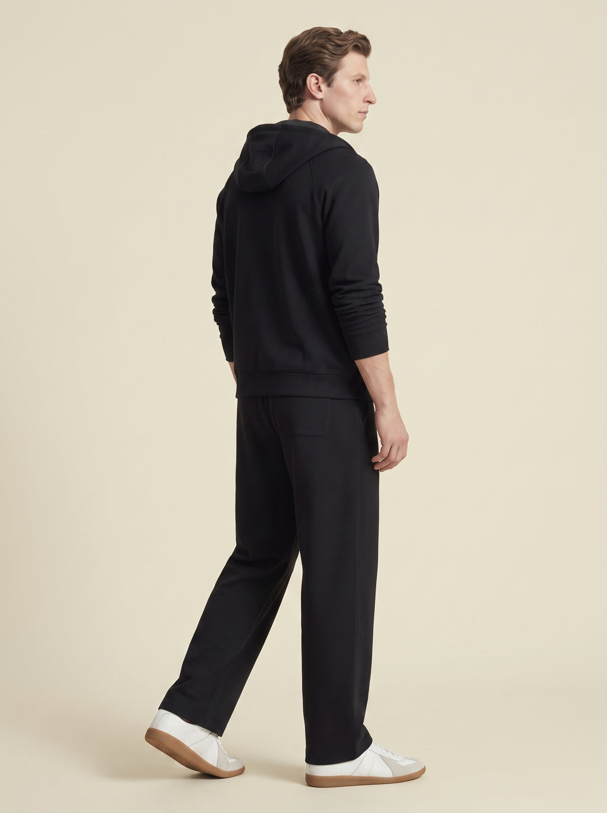 #Jet-Black_group A man wearing a black pullover hoodie and matching black trousers, shown from a side-back angle against a solid light beige studio background.