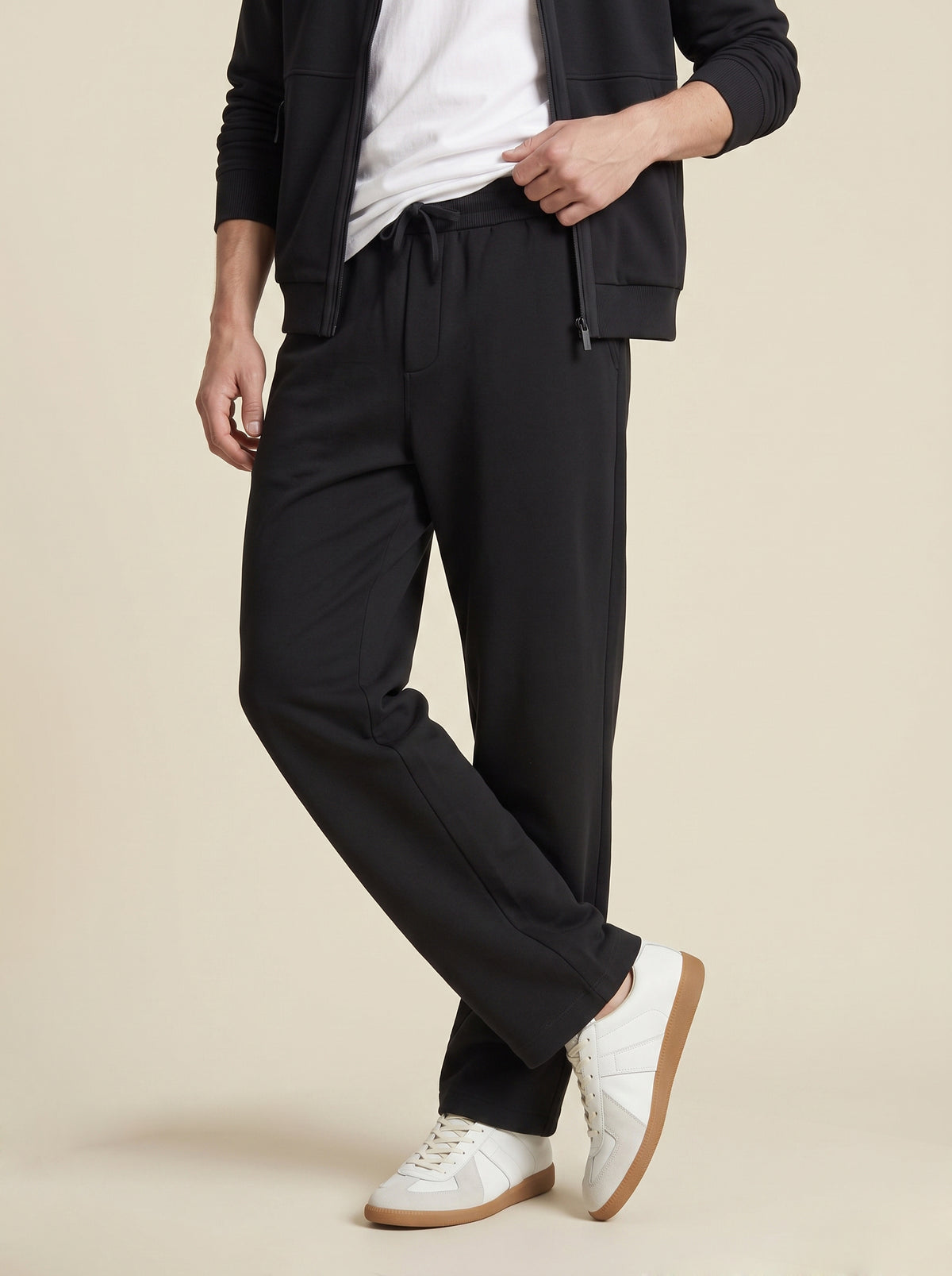 #Jet-Black_group A man wearing black drawstring trousers, styled with a white t-shirt, a black zip-up jacket, and white sneakers, standing against a solid beige background.