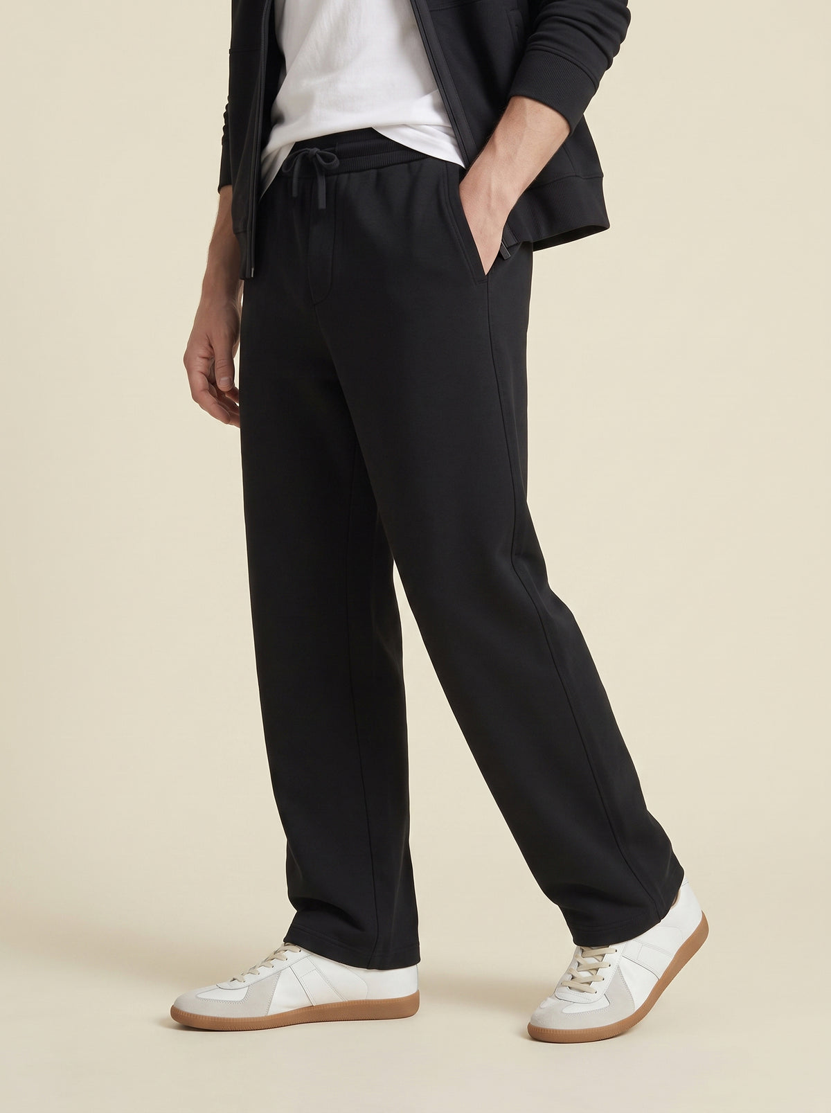 #Jet-Black_group A person wearing black drawstring trousers with a white t-shirt, black blazer, and white sneakers, standing against a solid beige background.