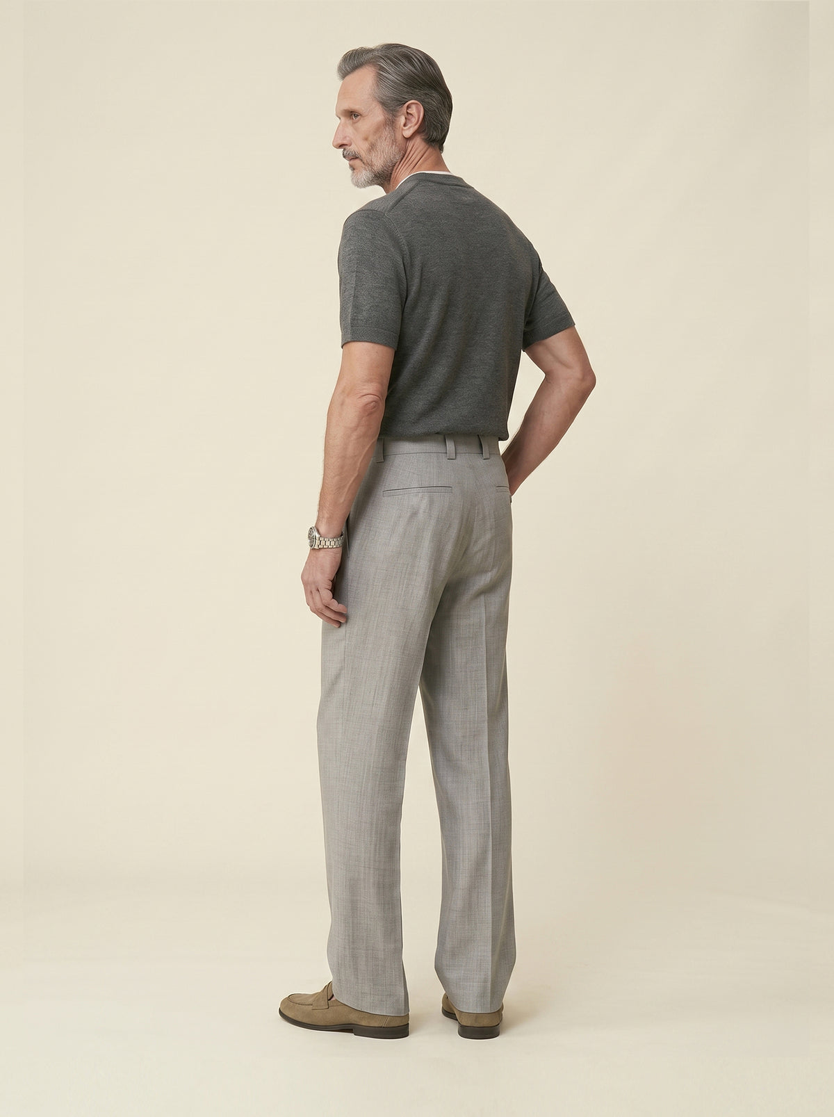 #Silver-Grey_group A man standing in profile wearing a dark gray short-sleeve knit polo shirt and light gray trousers, photographed against a plain warm beige studio background.