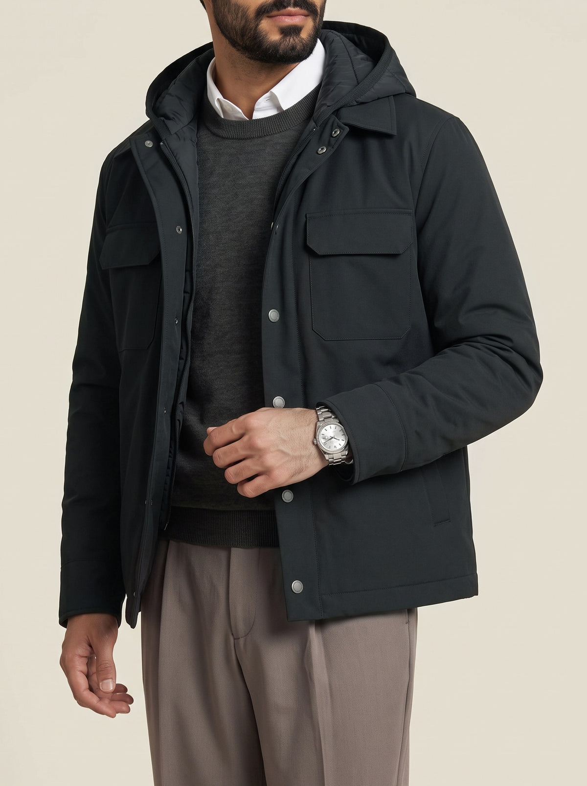 #Slate-Grey_group A man wearing a black hooded utility jacket over a dark gray sweater and white collared shirt, paired with taupe trousers, photographed against a plain light beige studio background.