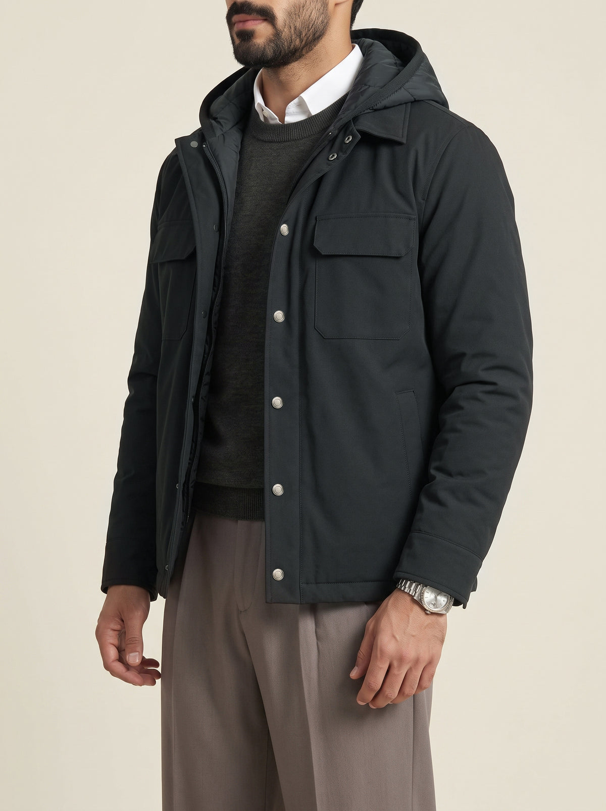 #Slate-Grey_group A man wearing a black hooded jacket over a dark sweater and white collared shirt, paired with taupe trousers, photographed against a plain light beige studio background.