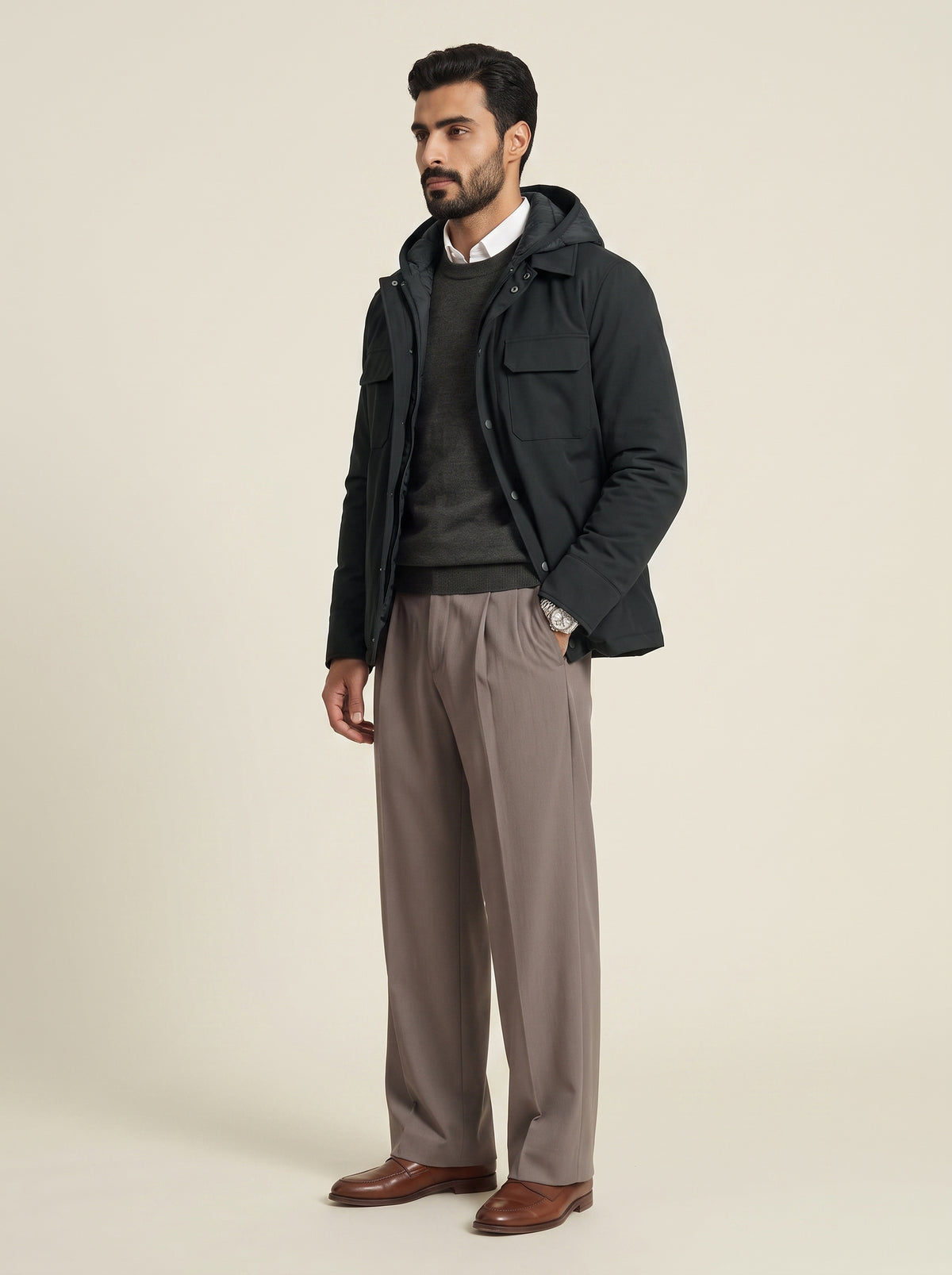 #Slate-Grey_group A man modeling a dark hooded jacket over a dark sweater, paired with taupe trousers and brown leather shoes, photographed against a plain light beige studio background.