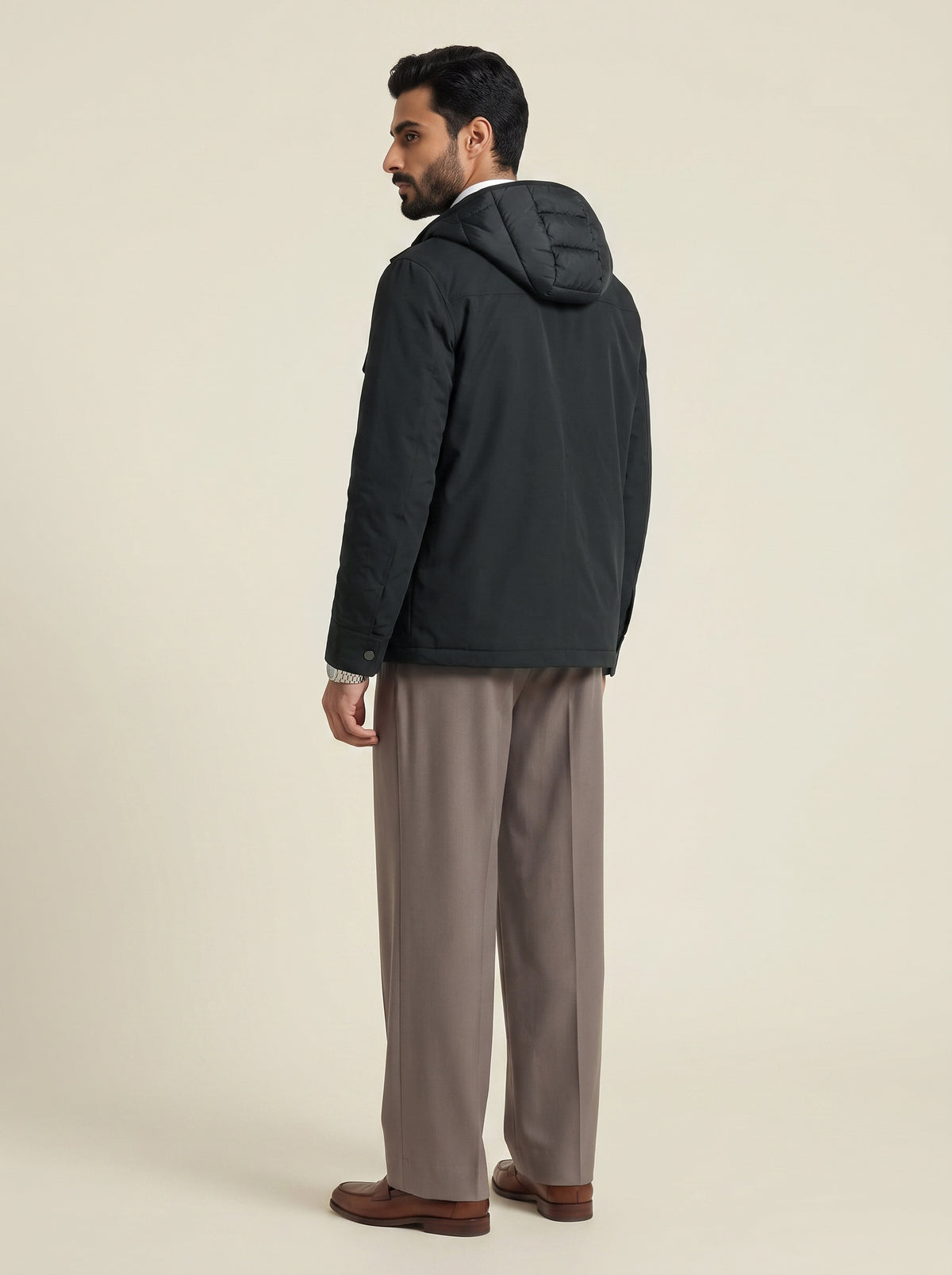 #Slate-Grey_group A man is shown from the back wearing a black hooded puffer jacket, taupe trousers, and brown leather shoes, standing against a solid light beige studio background.